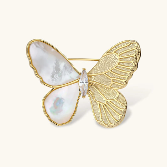 Gold-plated butterfly brooch with mother-of-pearl left wings, engraved gold right wings, and central zirconia crystal on cream background.
